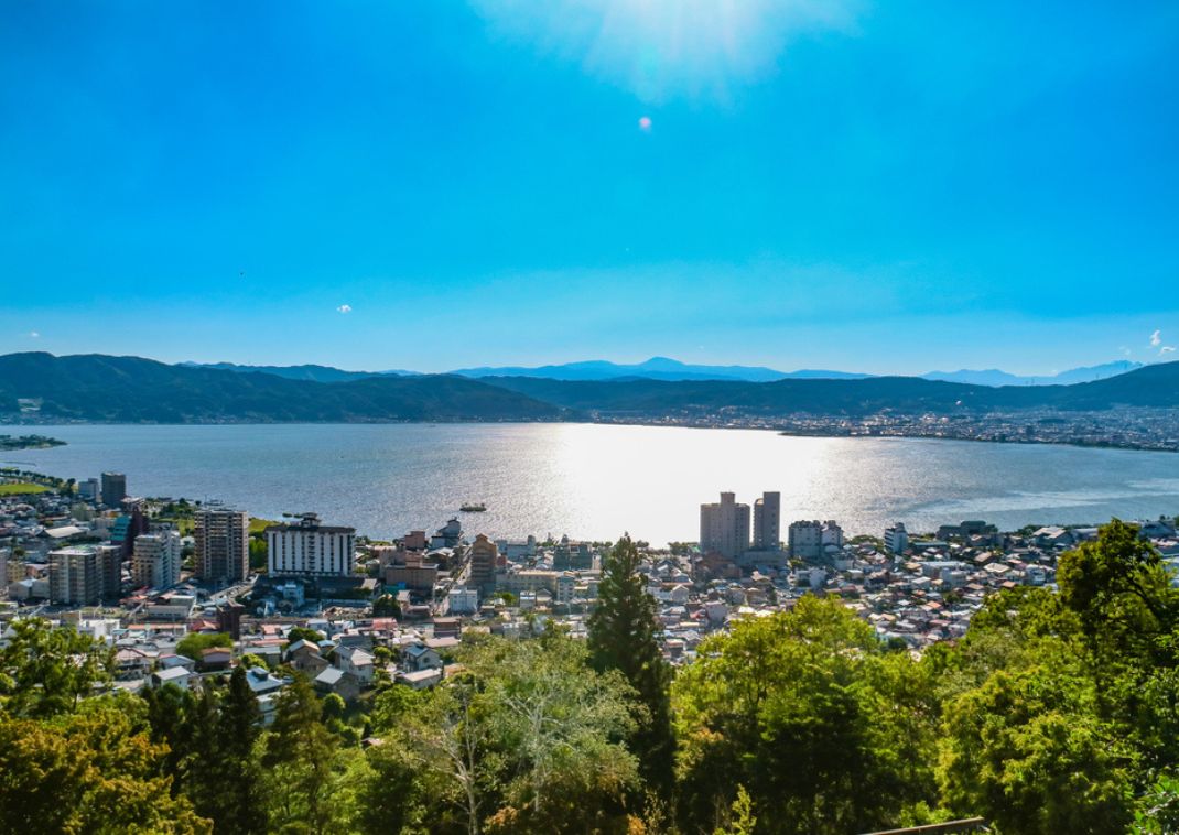 View of a large Japanese city by a sparkling lake, surrounded by mountains and forests under a bright blue sky