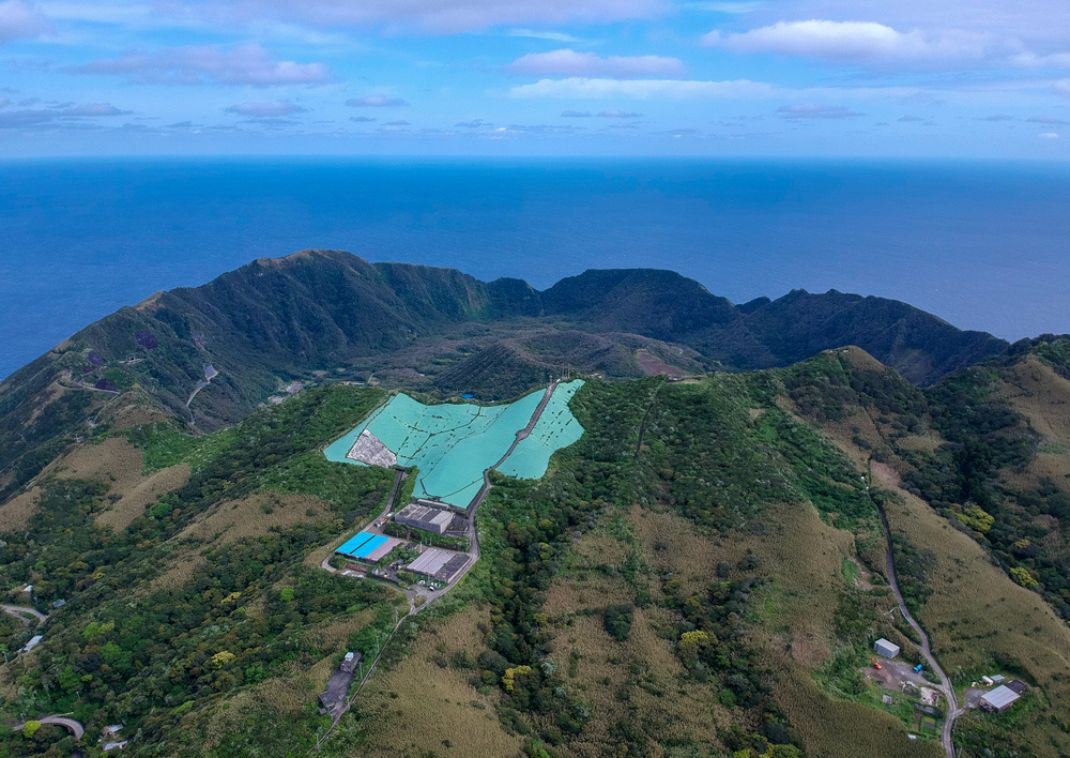 Aerial view of a lush mountainous landscape overlooking the ocean, with installations and agricultural greenhouses visible at the center of the image