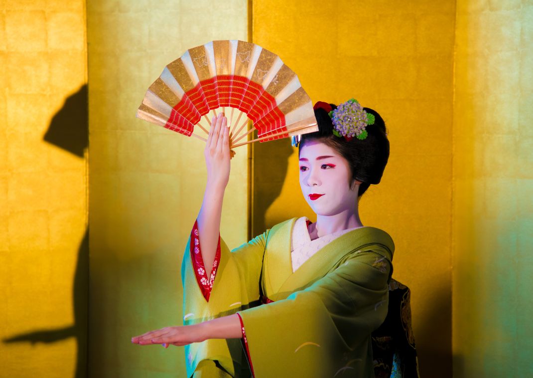 Japanese woman in a green kimono performing a traditional dance with a decorative folding fan against a gold background.