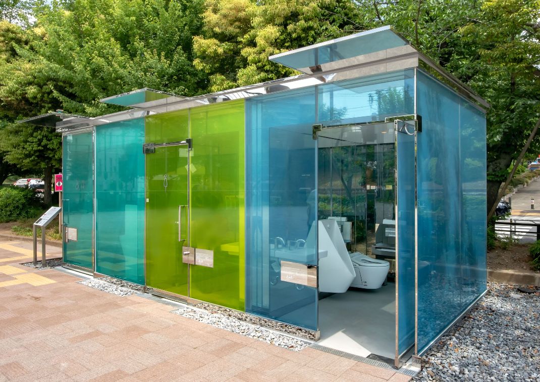 Transparent public toilets made of blue and green colored glass, with visible fixtures inside, located in a green outdoor setting.