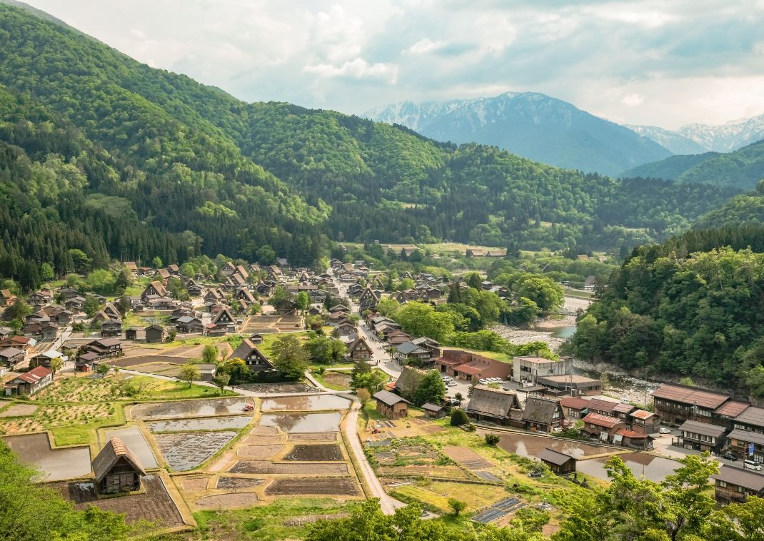 Panoramic view of Gokayama village in spring, with traditional thatched-roof houses, rice paddies, and lush green mountains in the background.