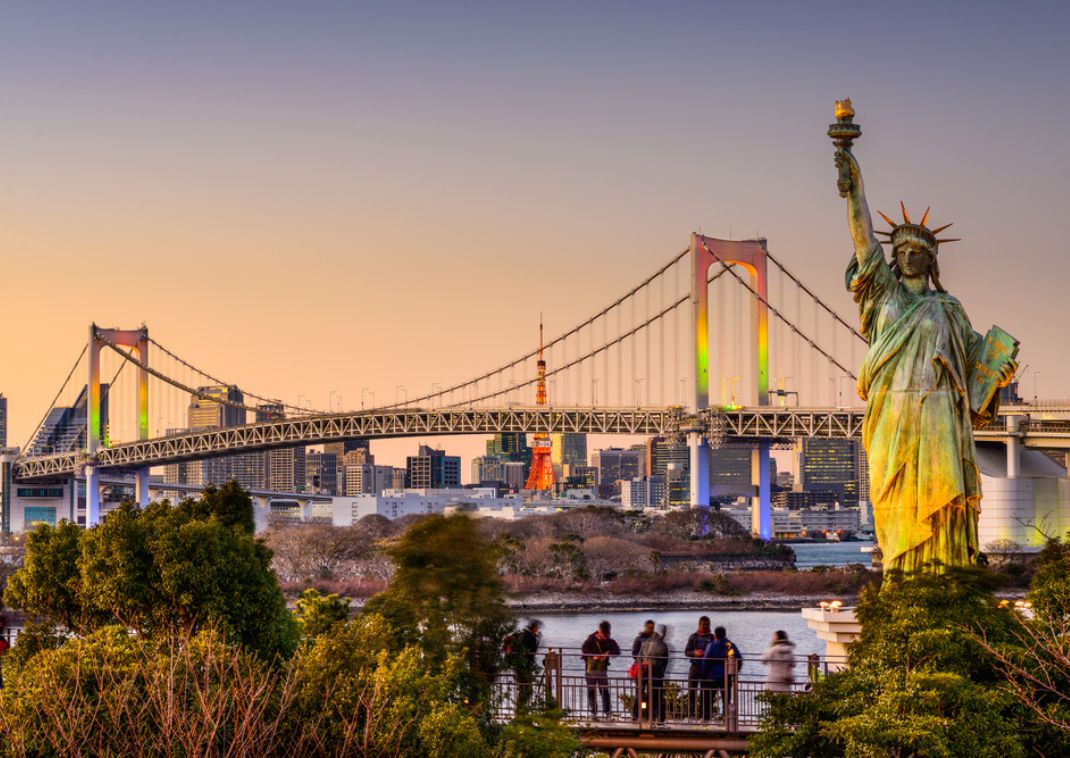 Replica of the Statue of Liberty in Odaiba, Tokyo, with the illuminated Rainbow Bridge and the Tokyo city skyline at sunset, including Tokyo Tower in the background.