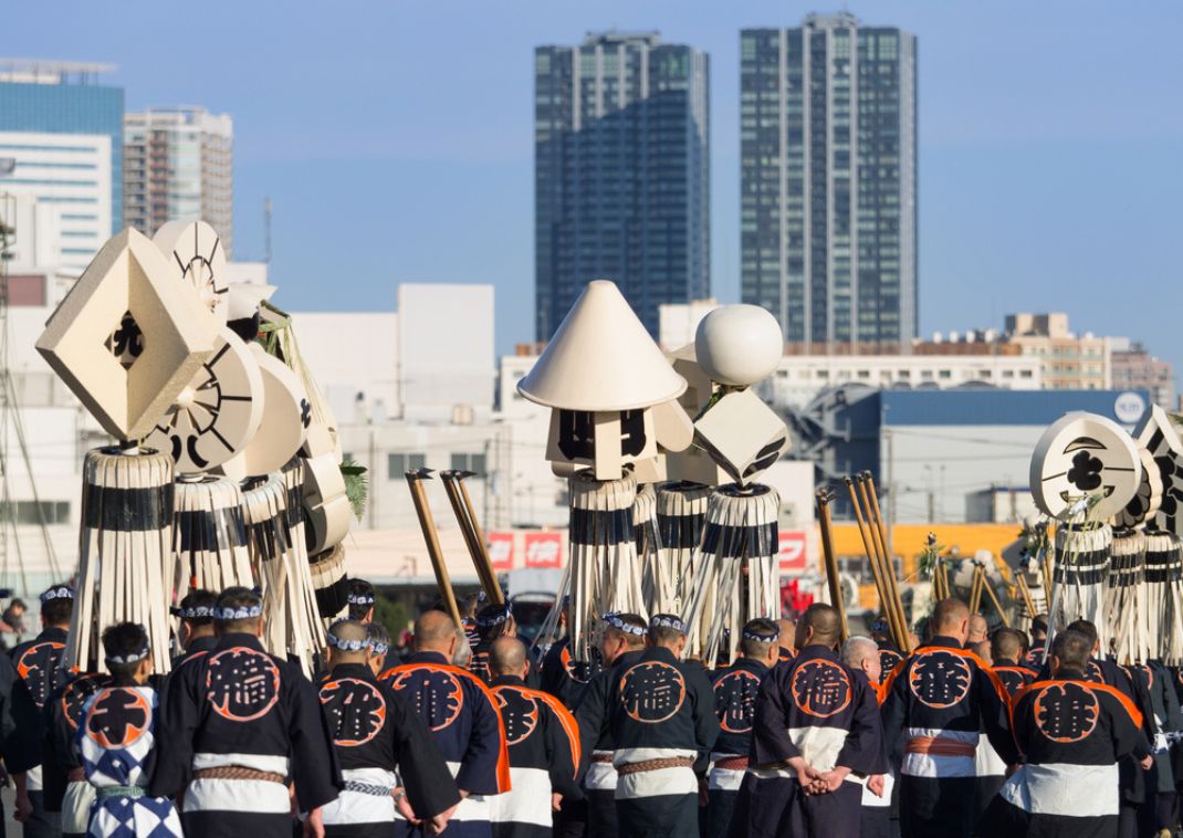 Group of participants in traditional Japanese festival attire carrying large decorative standards and lanterns, with modern Tokyo skyscrapers in the background under a clear blue sky