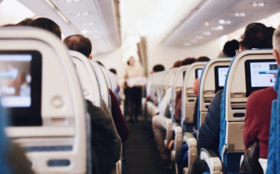 Interior of a large passenger airplane with people seated and a stewardess in uniform walking down the aisle