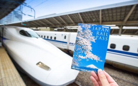 Hand holding a Japan Rail Pass ticket in front of a Shinkansen at Tokyo Station