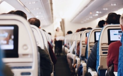 Interior of a large passenger airplane with people seated and a stewardess in uniform walking down the aisle