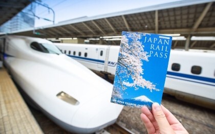 Hand holding a Japan Rail Pass ticket in front of a Shinkansen at Tokyo Station