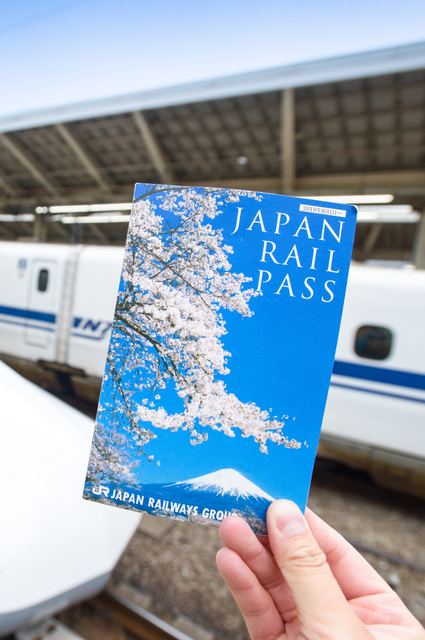 Hand holding a Japan Rail Pass ticket in front of a Shinkansen at Tokyo Station
